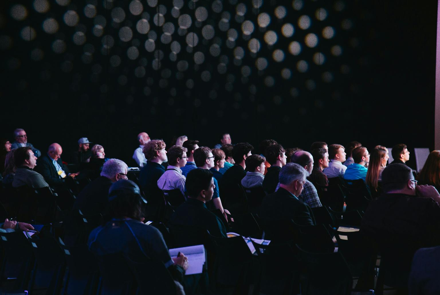 An audience seated in a dark auditorium or theater, with people in business attire facing a stage or presentation.