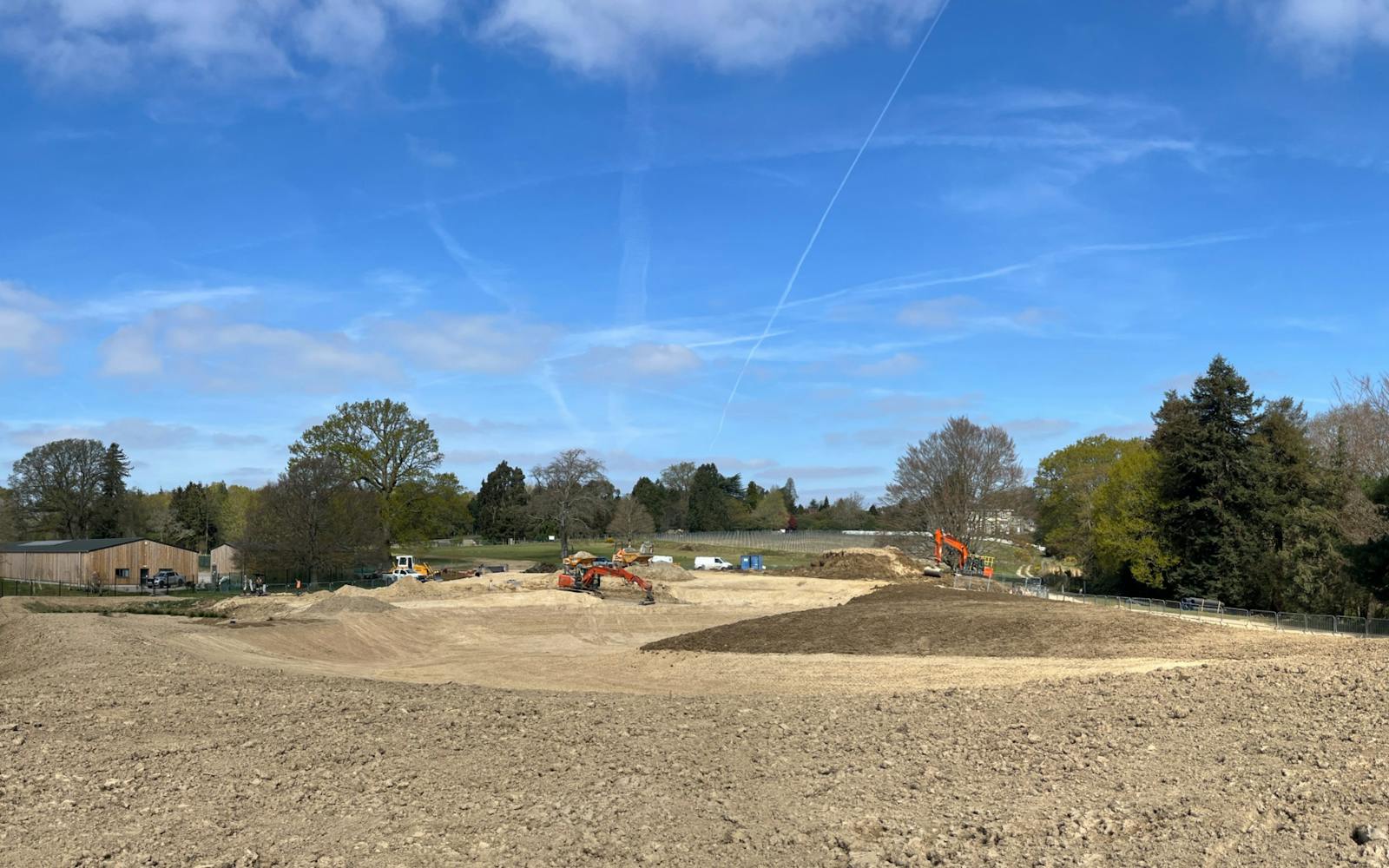 The image shows a construction site with a large expanse of bare earth in the foreground. Several pieces of heavy machinery, including excavators, are visible in the middle ground. The background features trees and a building, all under a blue sky with scattered clouds.