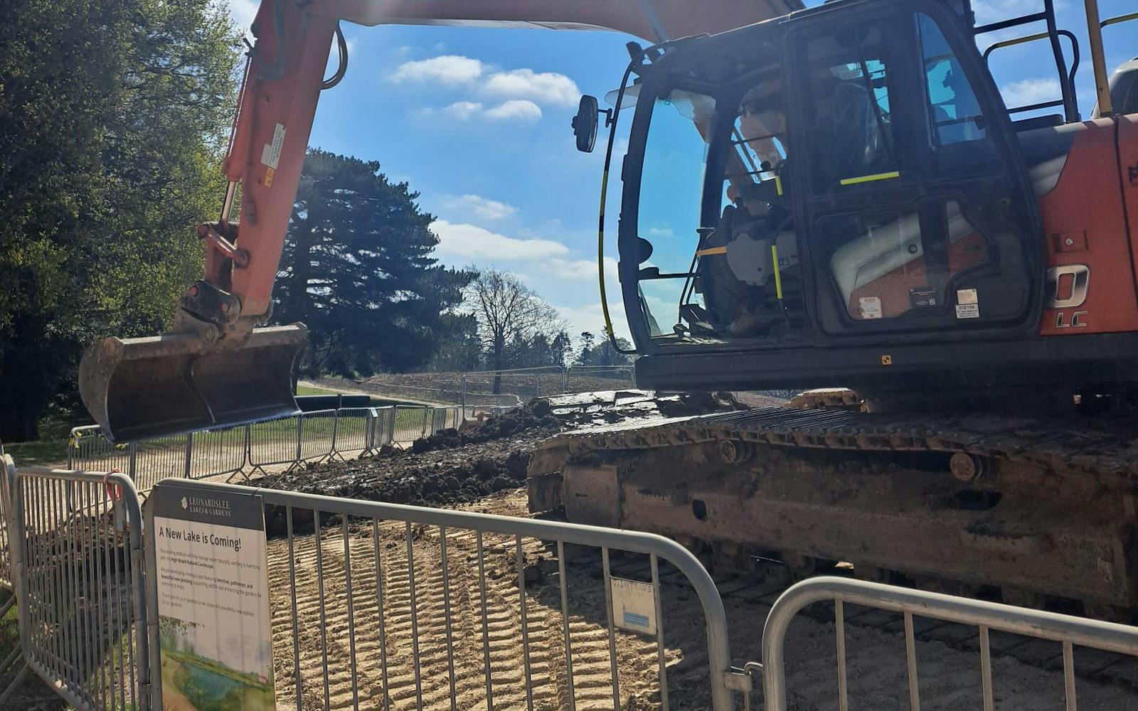 The image shows a construction site with an orange excavator in the foreground. In the background, there are trees, a body of water, and a blue sky with the sun shining. A metal fence is visible in the foreground, with a sign attached. The sign mentions a new lake and landscape.