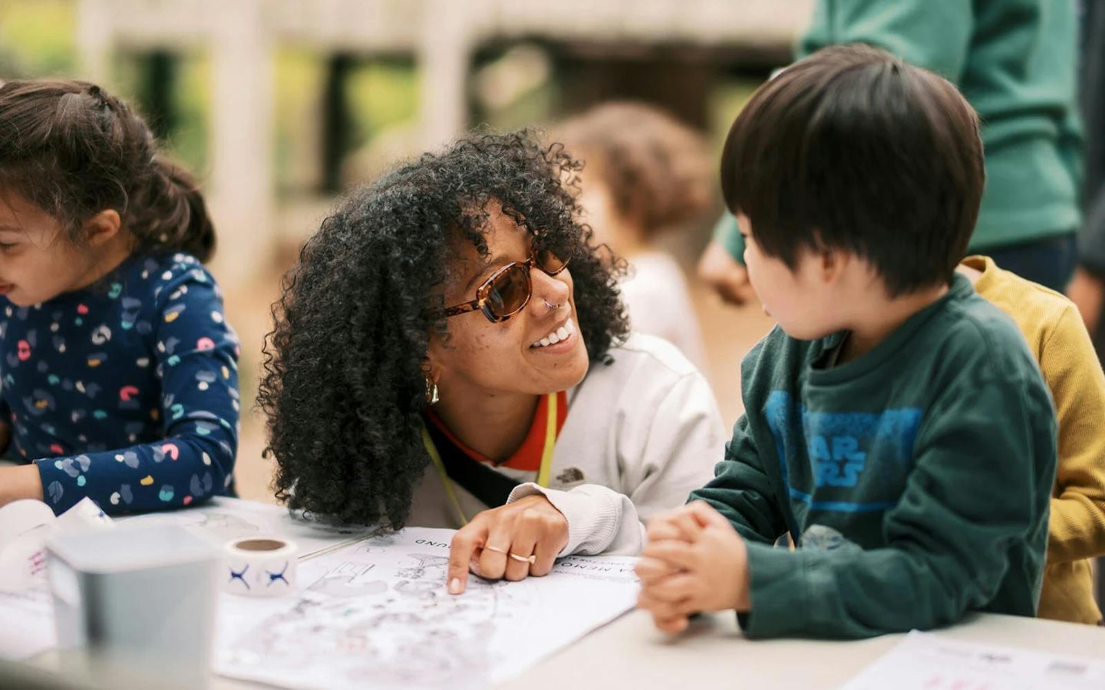 A woman smiles and talks with a young boy at a table covered in colouring sheets.