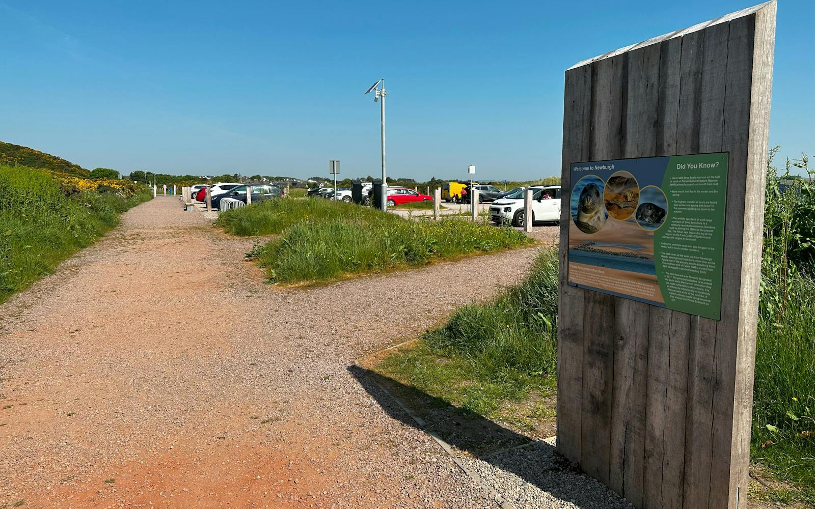 A sign next to a car park with blue skies and a path nearby