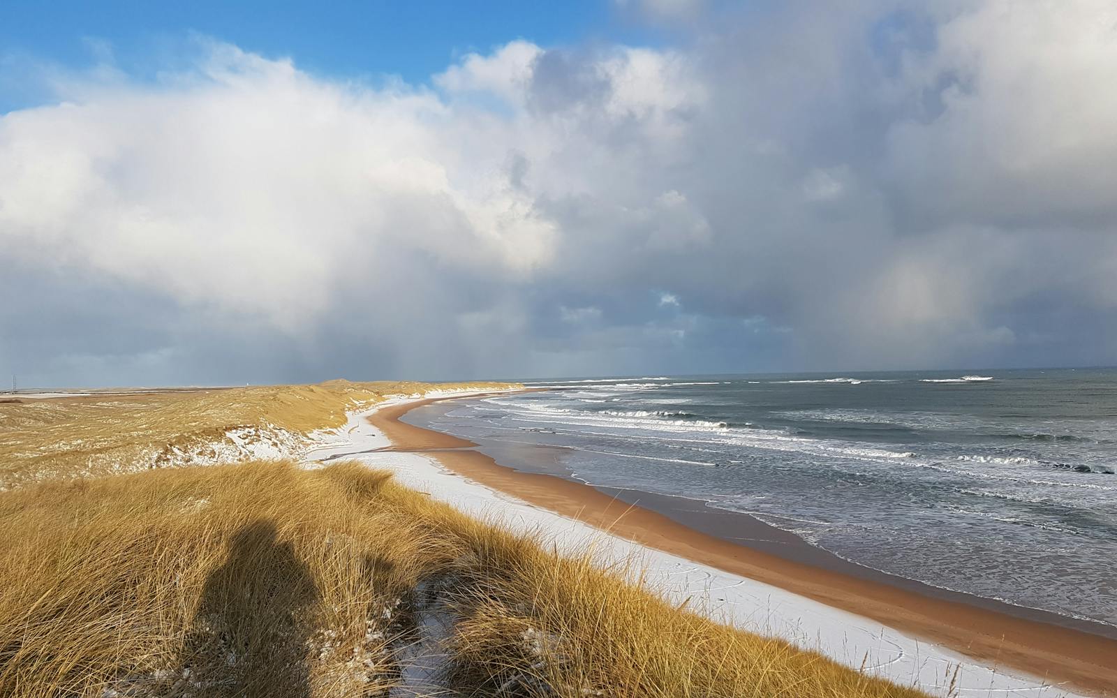 A landscape photograph shows a sandy and snowy beach, under a cloudy sky.