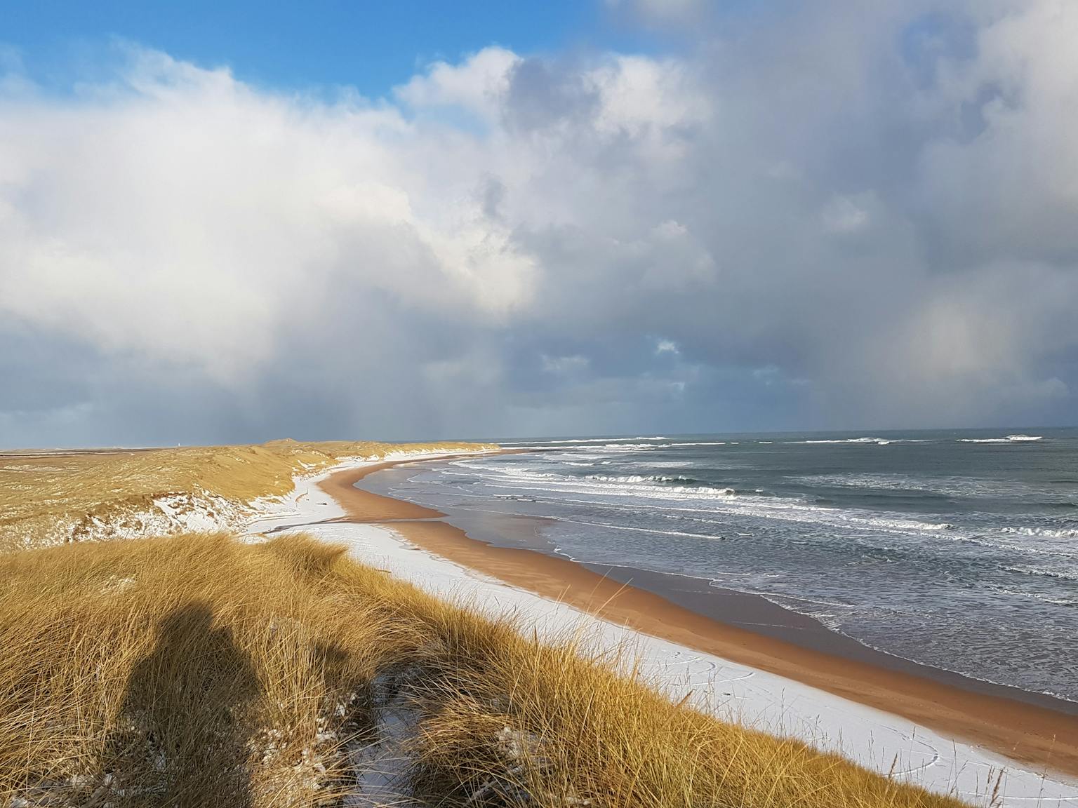 A landscape photograph shows a sandy and snowy beach, under a cloudy sky.