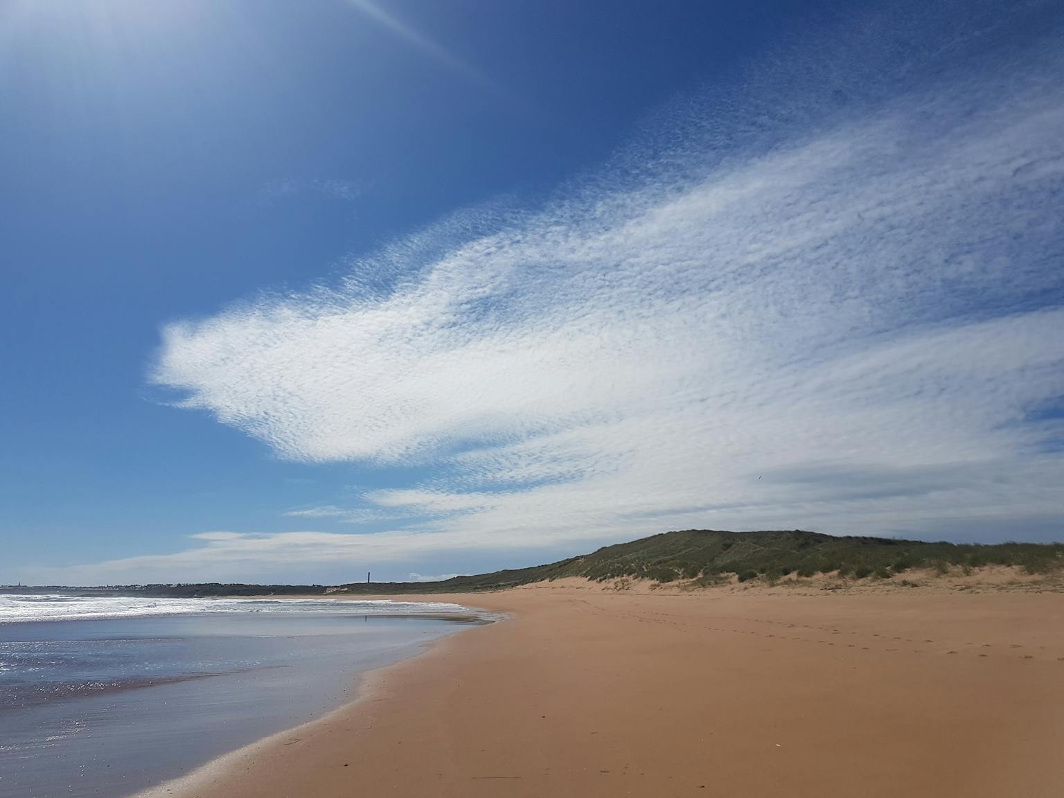A wide-angle landscape photograph shows a sandy beach, with hills in the background, under a blue and cloudy sky.