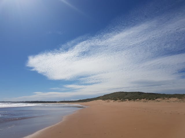 A wide-angle landscape photograph shows a sandy beach, with hills in the background, under a blue and cloudy sky.