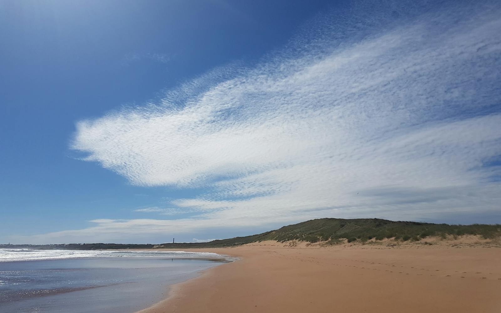 A wide-angle landscape photograph shows a sandy beach, with hills in the background, under a blue and cloudy sky.