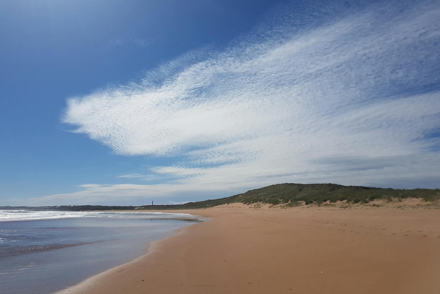 A wide-angle landscape photograph shows a sandy beach, with hills in the background, under a blue and cloudy sky.