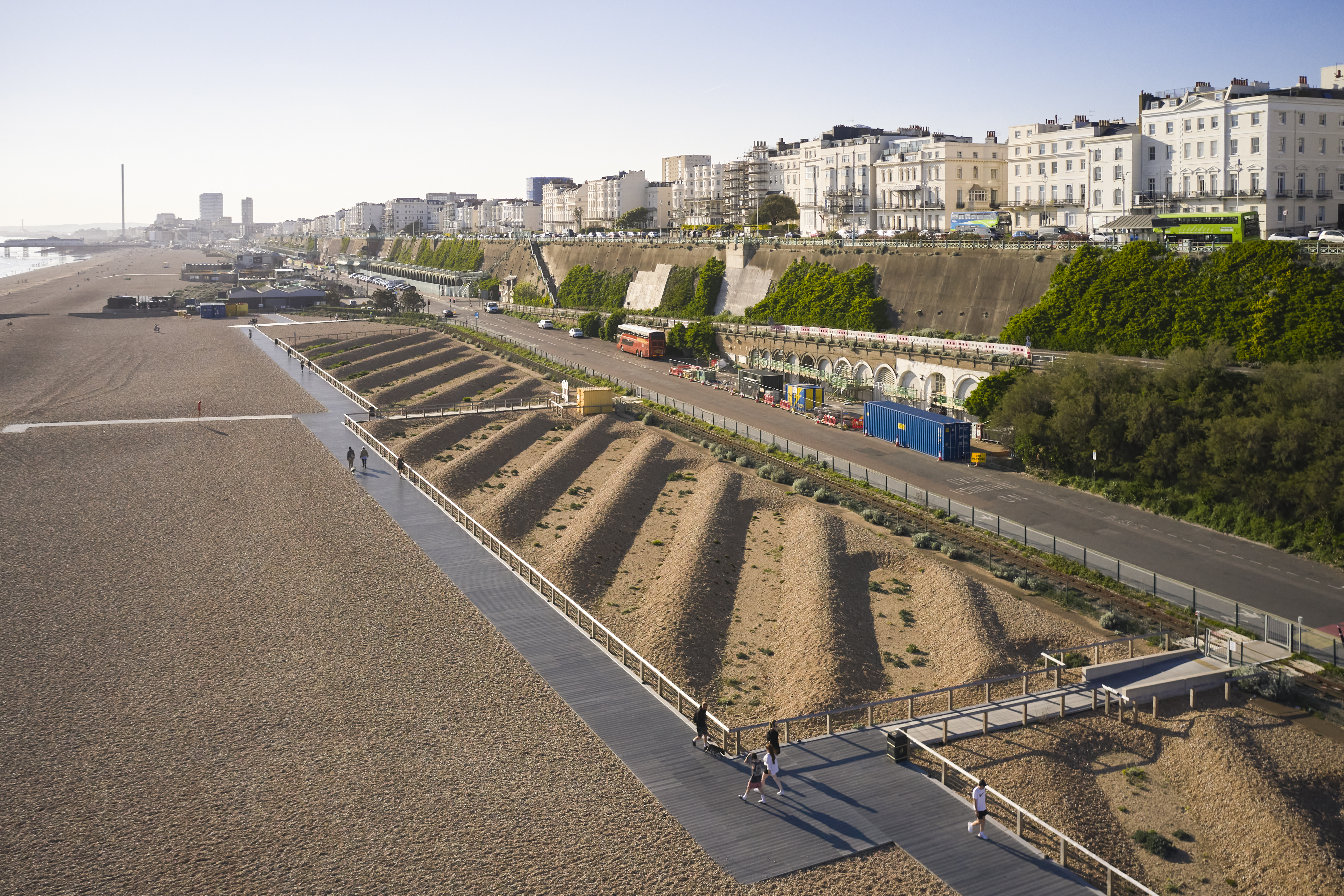 A sandy beach and boardwalk next to a road, with white buildings and a cliff in the background.