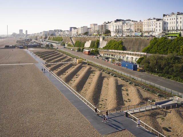 A sandy beach and boardwalk next to a road, with white buildings and a cliff in the background.