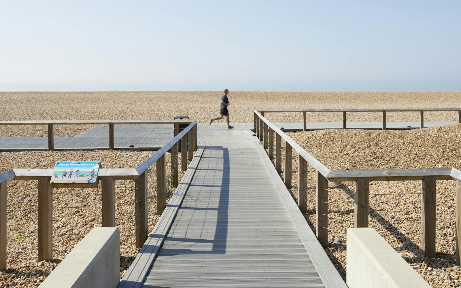 A person jogs along a boardwalk on a pebble beach under a clear sky.