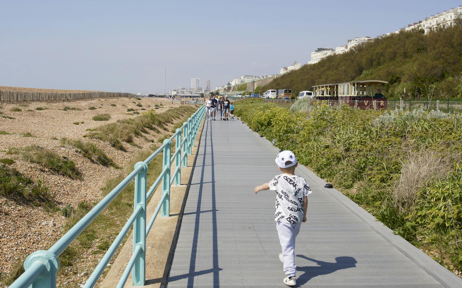 A person in a white shirt and hat walks along a boardwalk next to a beach and grassy hill.