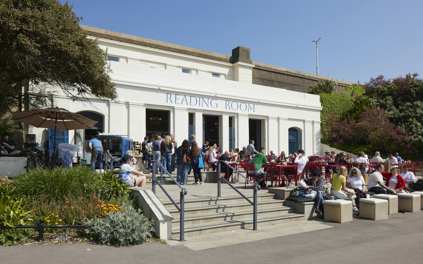 A white building with "READING ROOM" written on it, with people sitting outside at tables and on benches.