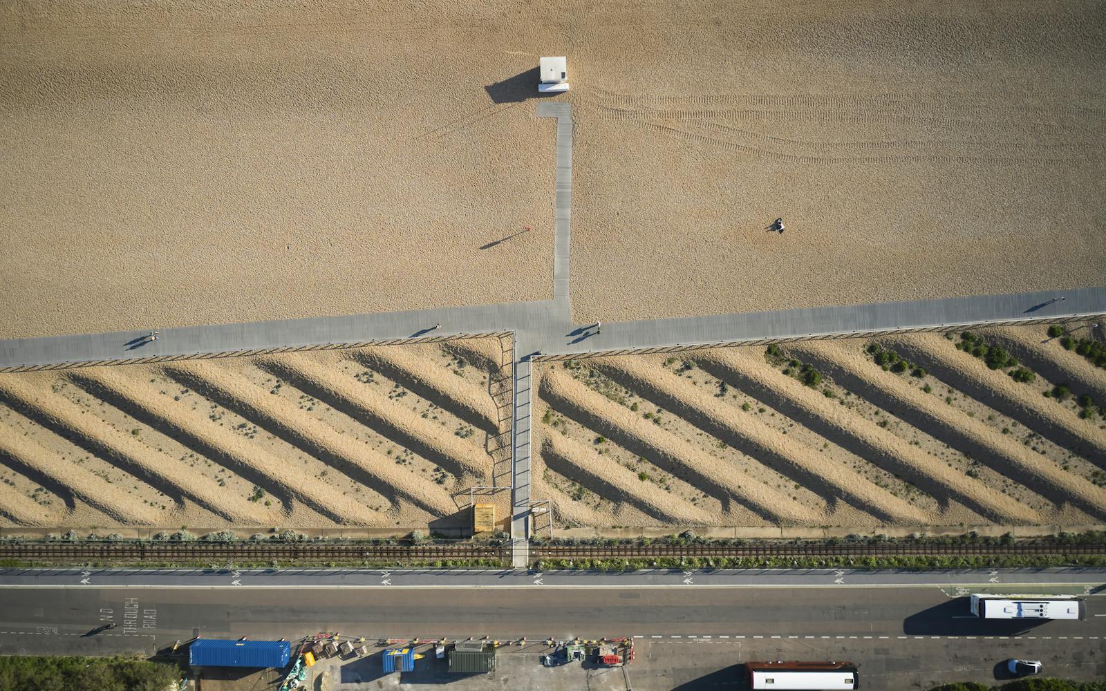An aerial view of a sandy beach with a small building, a boardwalk, and a road with vehicles.