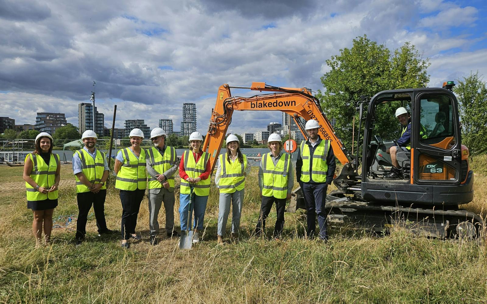 A group of eight adults wearing hard hats and high-visibility vests stand on a grassy patch of ground with an orange excavator behind them. One person is operating the excavator. In the background, modern buildings are visible under a cloudy sky.