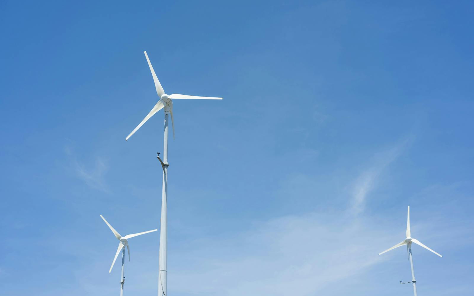A low-angle wide shot of three white wind turbines against a clear blue sky. The central turbine is the tallest, with two smaller turbines on either side, one to the lower left and one to the lower right. All turbines have three blades.