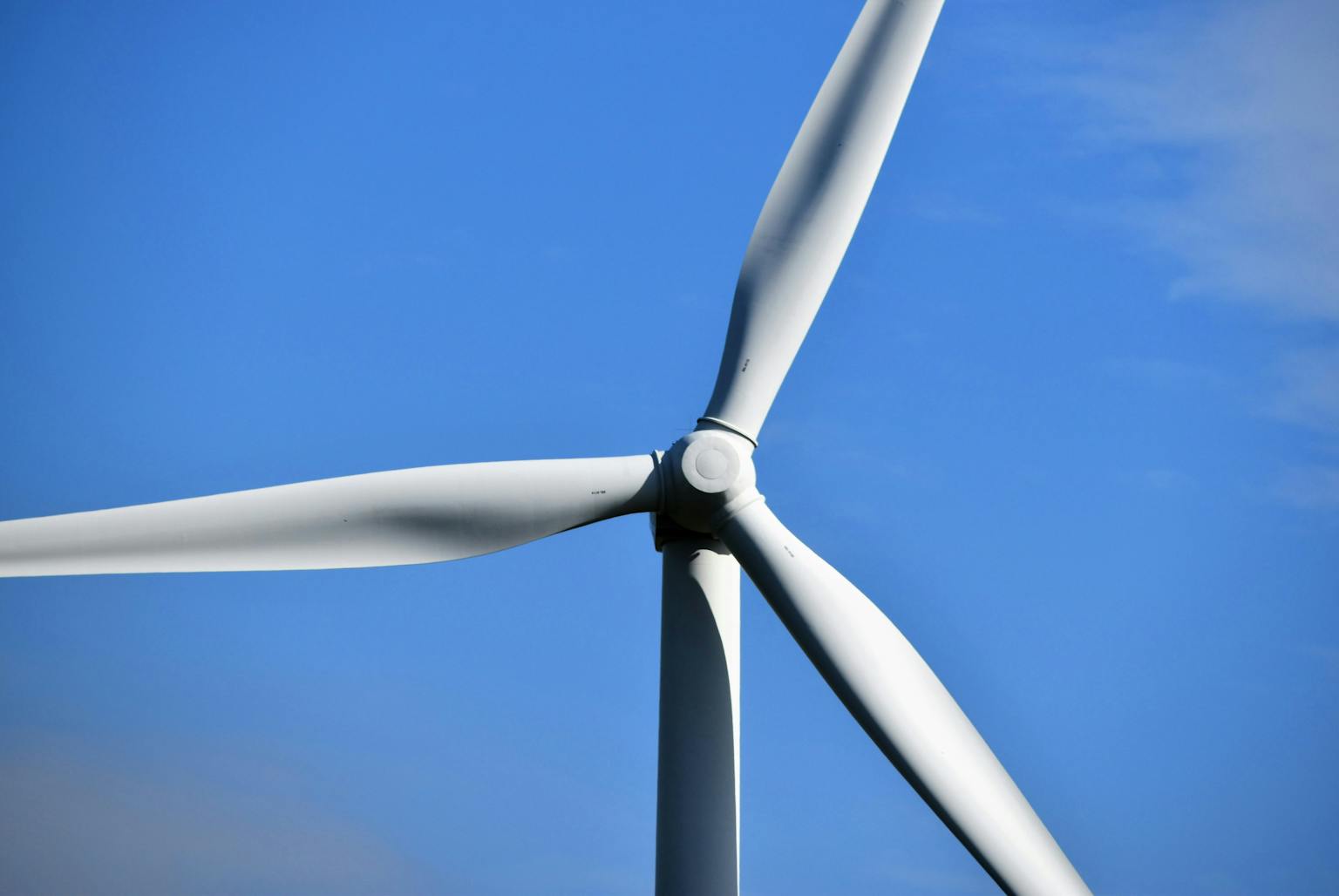 A close-up, low-angle shot of the top half of a white wind turbine against a clear blue sky, with its three blades prominently displayed.