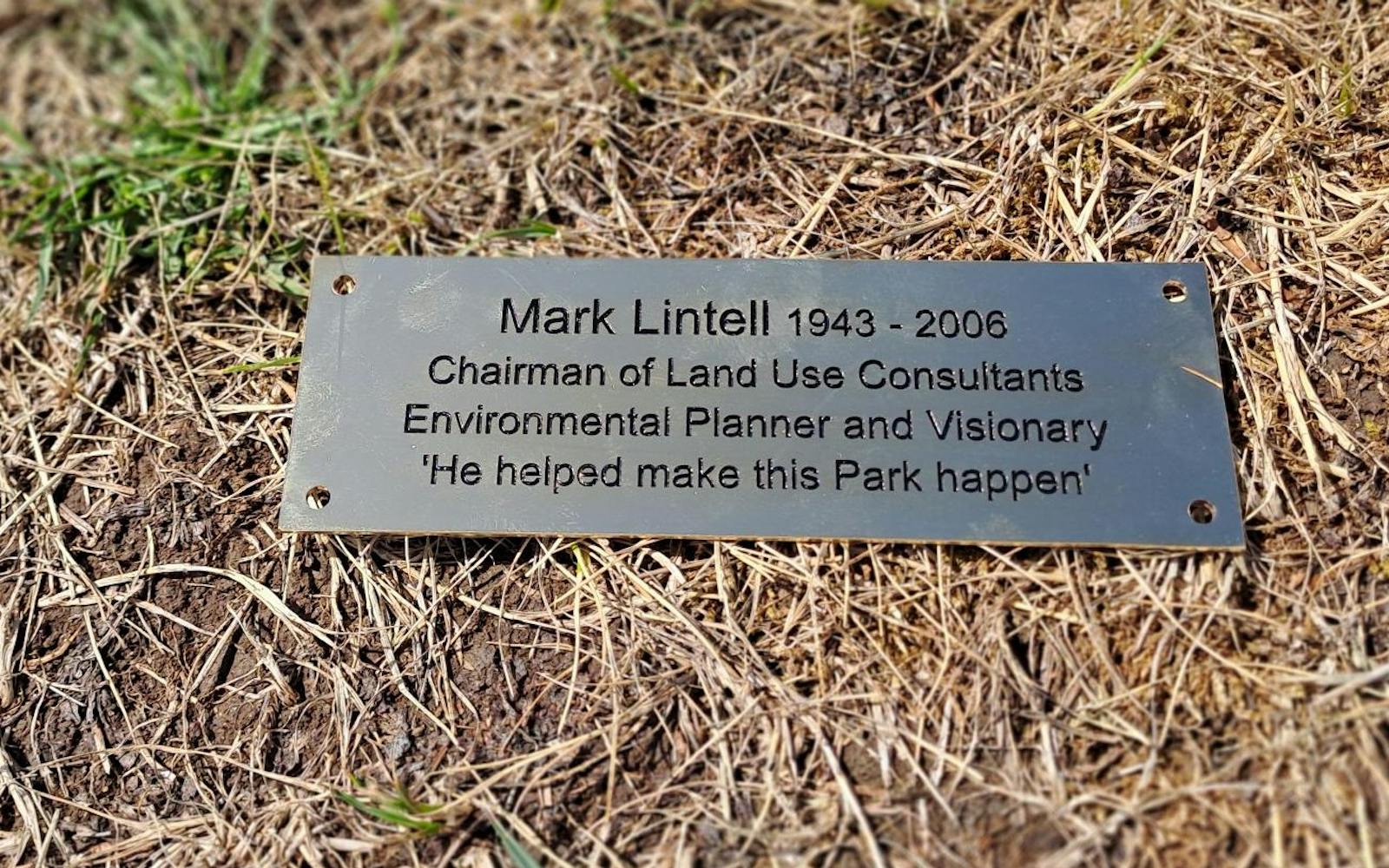 A close-up of a bronze plaque on the ground. It reads: "Mark Lintall 1943 - 2006 Chairman of Land Use Consultants Environmental Planner and Visionary 'He helped make this Park happen'". The plaque is surrounded by dry grass and some green blades.