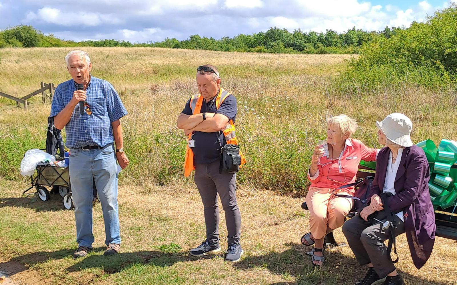A group of four adults are outdoors on a sunny day. On the left, a man in a blue plaid shirt and jeans is speaking into a microphone. Next to him, a man in an orange reflective vest and dark clothing stands with his arms crossed. To the right, two women are seated on a bench; one in a peach-colored top is holding a drink, and the other in a dark jacket and sun hat is looking towards the speaker. In the background, there is a field of dry grass and green trees under a partly cloudy sky.