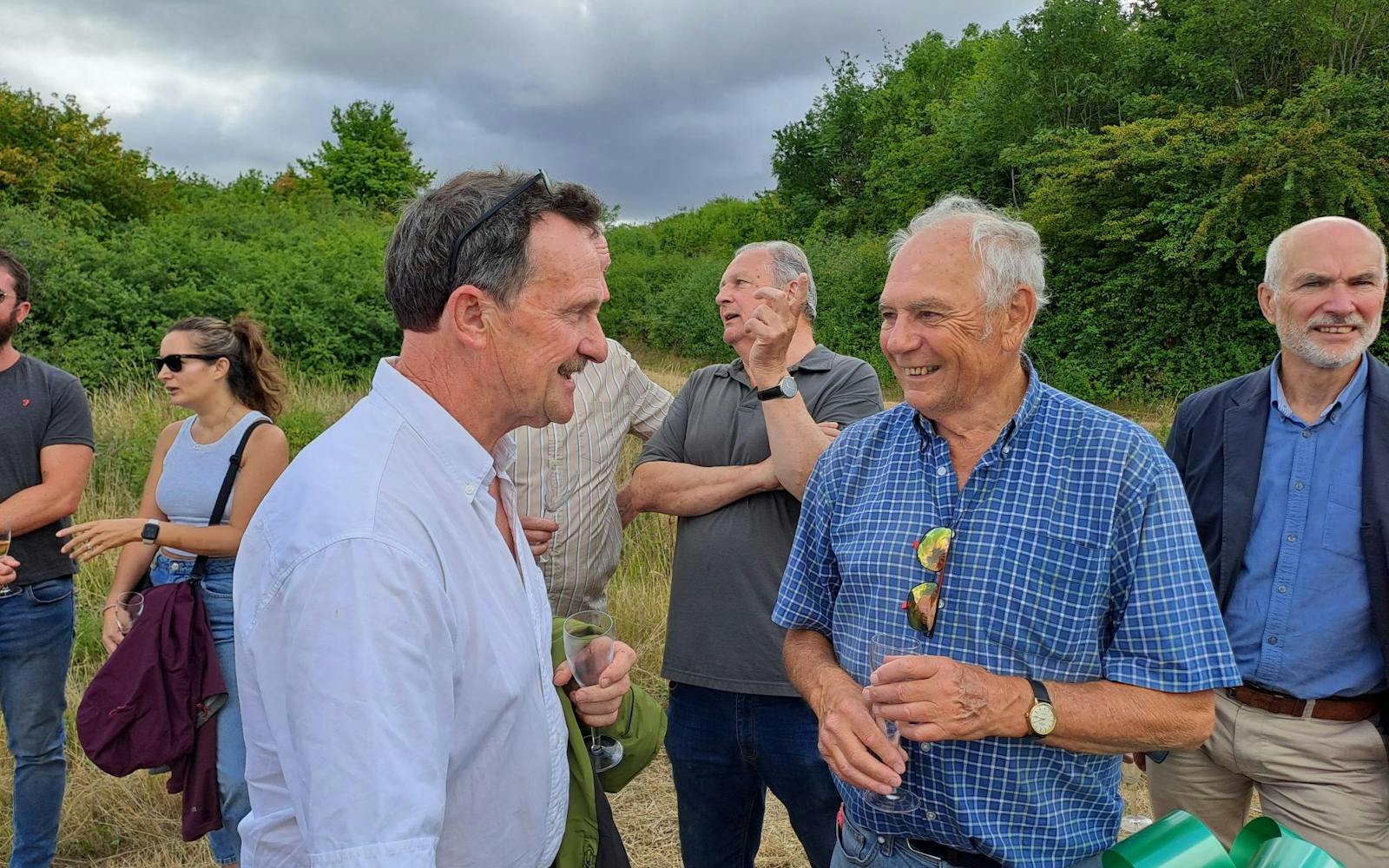 A group of adults are gathered outdoors on what appears to be a cloudy day. In the foreground, two men are facing each other and smiling, seemingly in conversation. The man on the left is wearing a white collared shirt, and the man on the right is wearing a blue plaid shirt with sunglasses hanging from his collar. Other individuals are visible in the background, including a woman in a light blue top and sunglasses, and another man with his hand raised to his face. Beyond the people, there's a natural landscape with trees and dry grass under a grey sky.