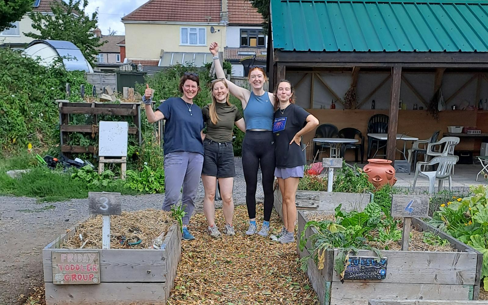 Four people are standing in a community garden, smiling and gesturing at the camera. Raised garden beds are in the foreground, and a building with a green roof is in the background.