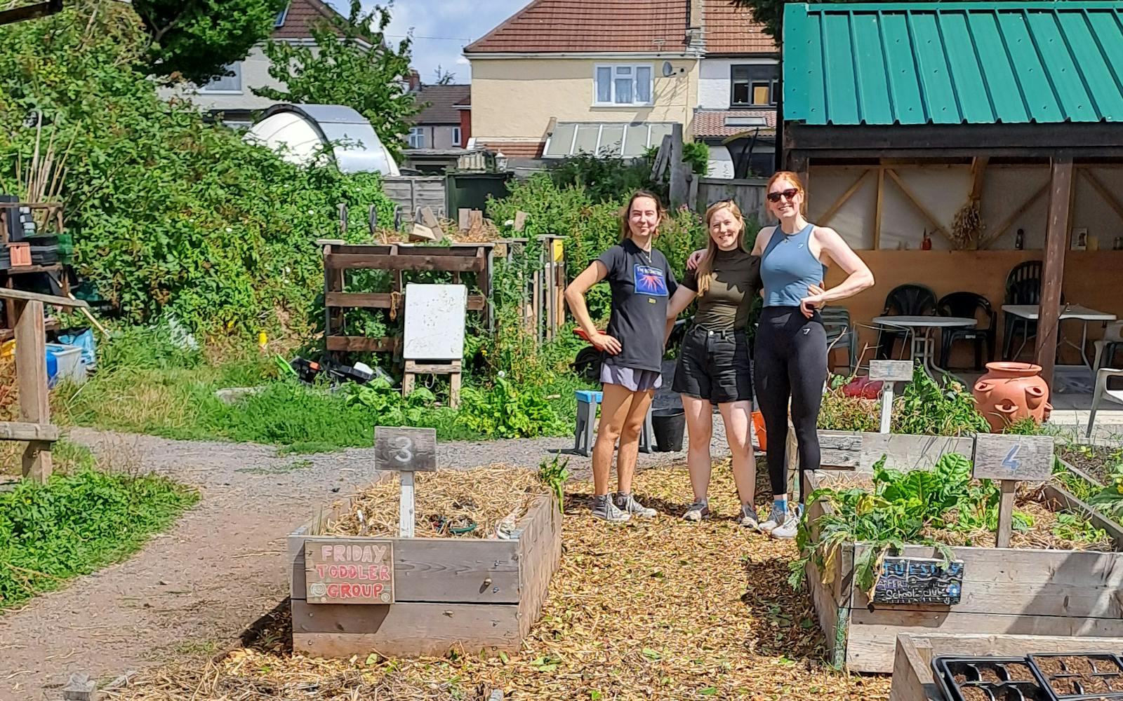 Three people are standing in a community garden, smiling at the camera. Raised garden beds are in the foreground, and a building with a green roof is in the background.