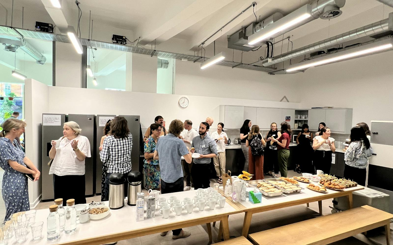 A group of people gathers in a modern, well-lit room, possibly an office kitchen or break room. There are long white tables laden with refreshments, including pastries, drinks in carafes, and stacks of mugs. People are standing in small groups, chatting and holding cups. The room has industrial-style ceiling lights and large stainless-steel refrigerators lining one wall.