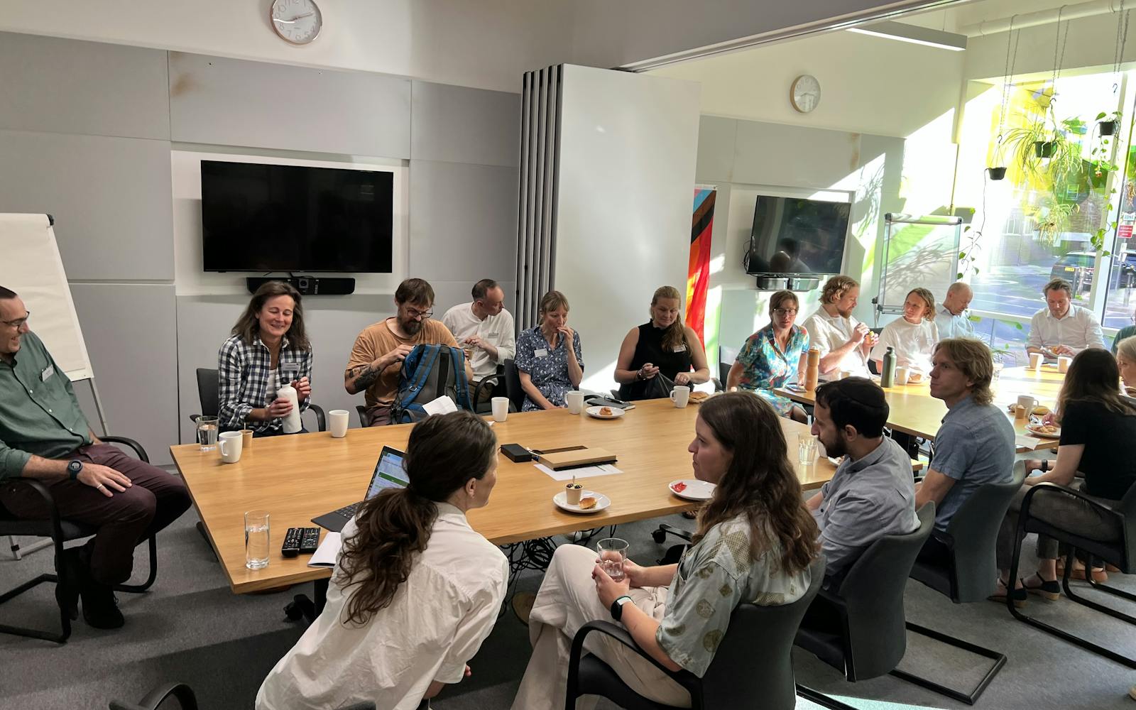 A diverse group of individuals sits around a large, light-colored wooden conference table in a modern meeting room. There are people on all sides of the table, some with laptops open, while others are engaged in conversation. Snacks and drinks are scattered on the table. The room has large windows on the right, allowing natural light to enter, and two flat-screen TVs are mounted on the wall behind the table. A whiteboard stands in the foreground to the left.