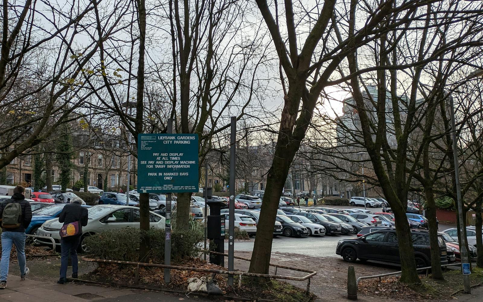 A city car park with numerous parked cars, viewed through leafless trees on a cloudy day. A green sign reads "Lushpark Gardens City Parking" with payment instructions. Two people with backpacks walk on the pavement nearby.