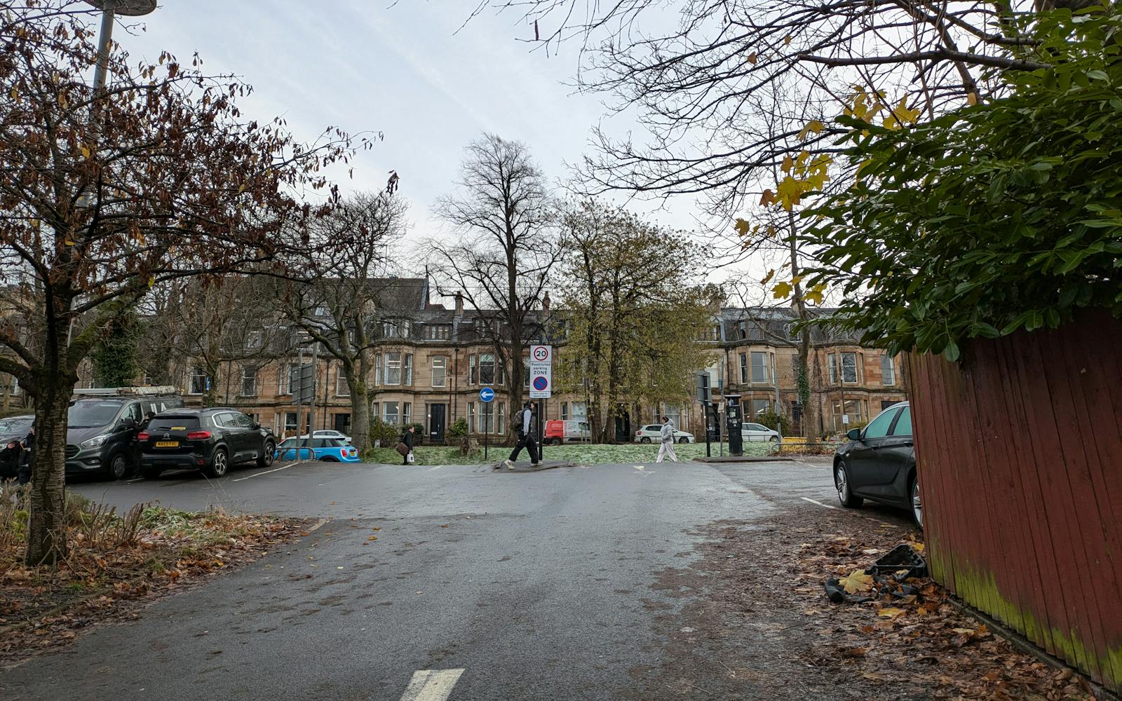 A quiet residential street in autumn, with wet pavement, fallen leaves, and parked cars. People walk near Victorian-style stone buildings in the background, surrounded by bare and leafy trees under an overcast sky.