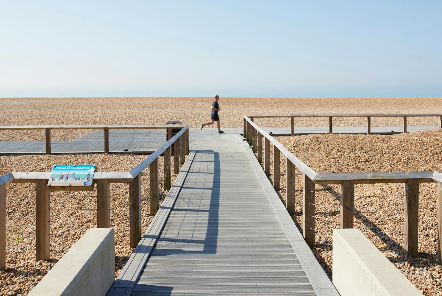 A wide shot of the revitalised Black Rock seafront in Brighton. It features a new grey boardwalk leading down to a pebble beach, with a person running in the distance under a clear blue sky.