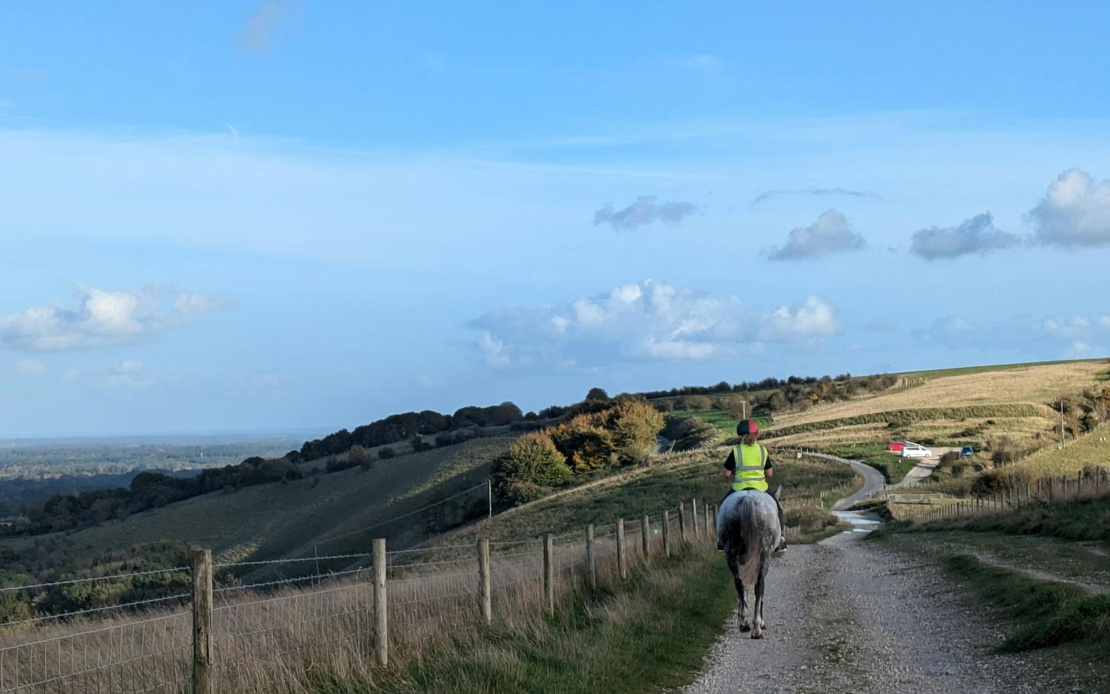 A person wearing a yellow high-visibility vest and a helmet is riding a gray horse along a dirt track. The path curves up a grassy hill with a wire and wooden fence running alongside it. In the background, there is a panoramic view of rolling hills and a valley under a partly cloudy blue sky.