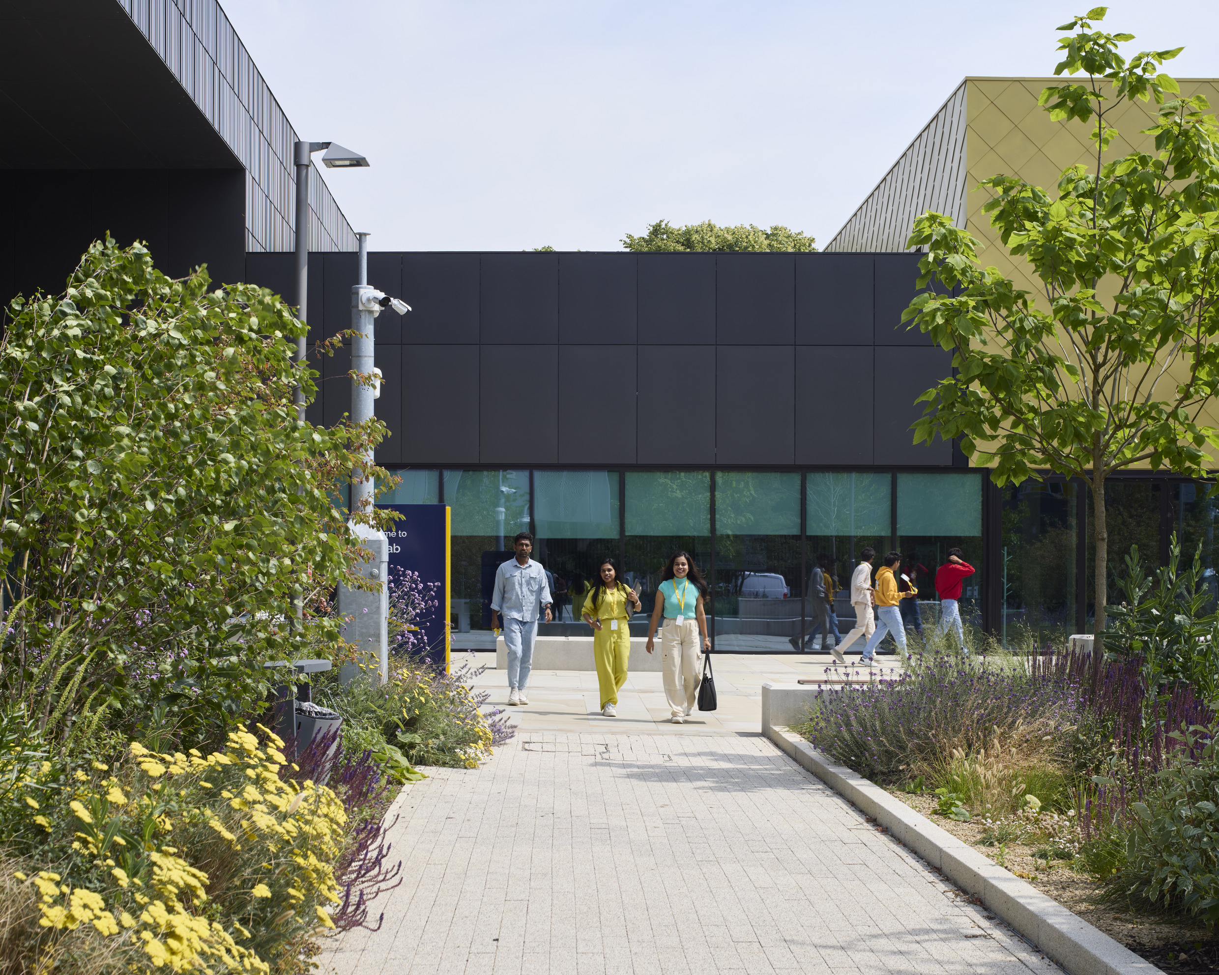 A group of people walk along a paved walkway between two modern university buildings with plants on either side.
