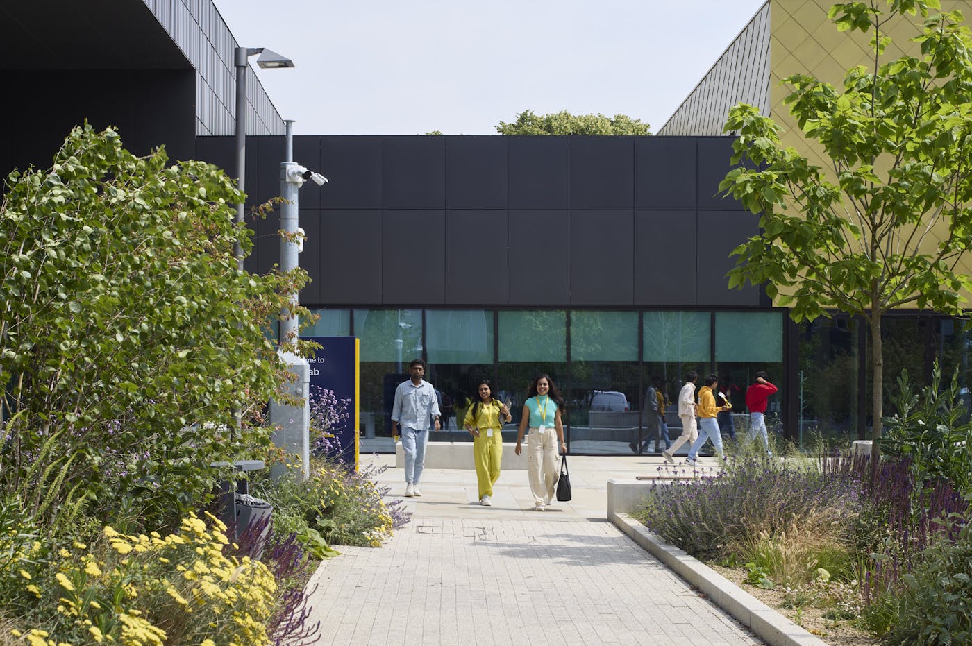 A group of people walk along a paved walkway between two modern university buildings with plants on either side.