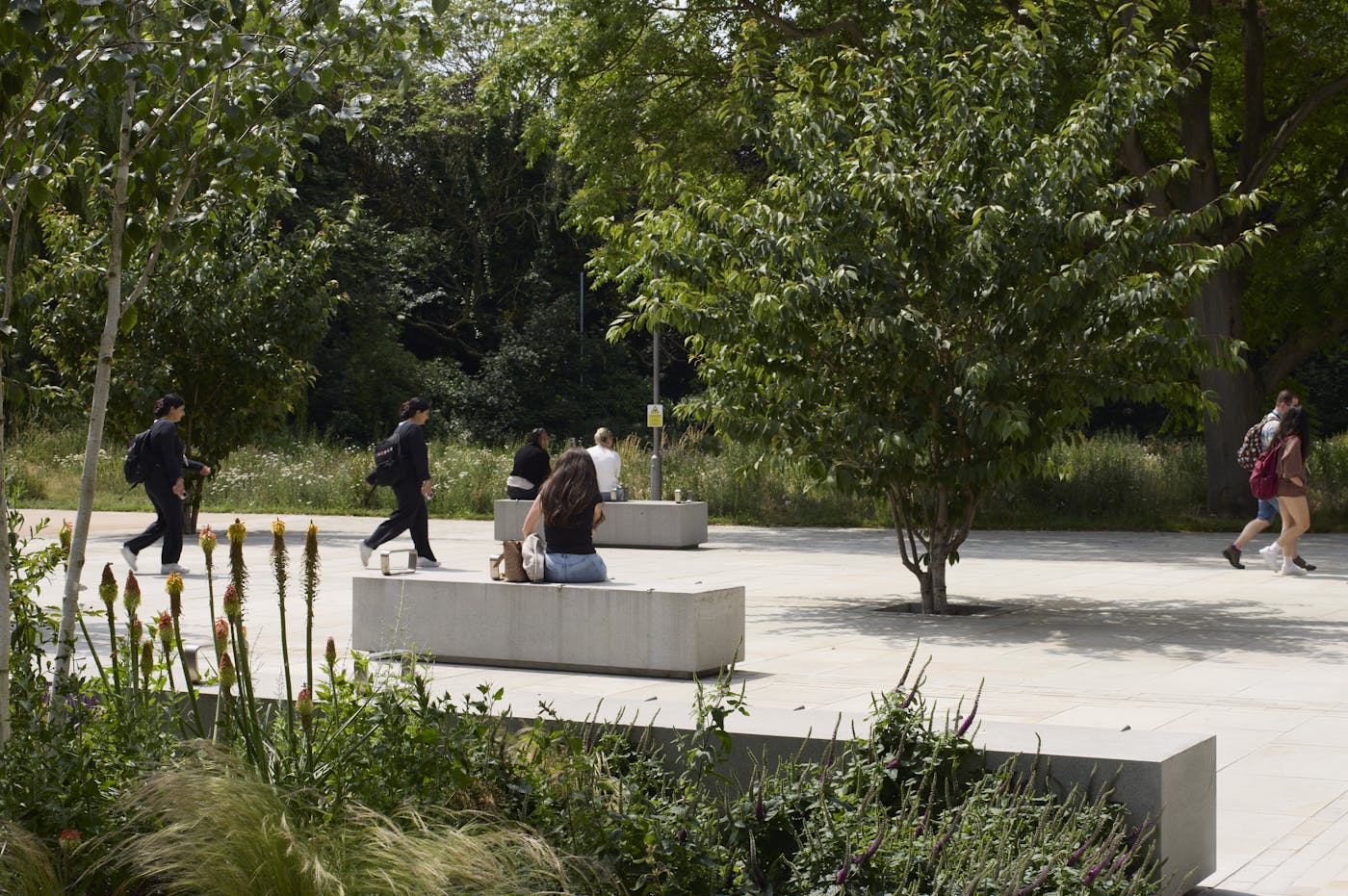 People are sitting on and walking by a stone bench in a leafy outdoor area.