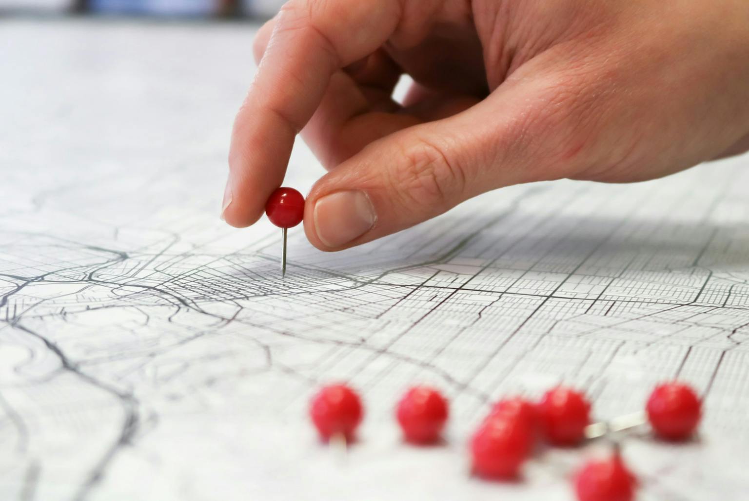 A close-up, high-angle shot of a person's hand placing a single red thumbtack on a paper map with a black and white street grid. There are several other red thumbtacks scattered in the foreground.