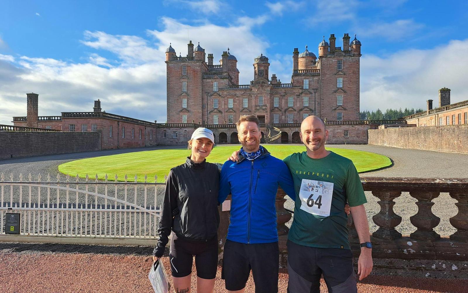 Three people wearing sports gear posing in front of a castle under a sunny and blue sky.