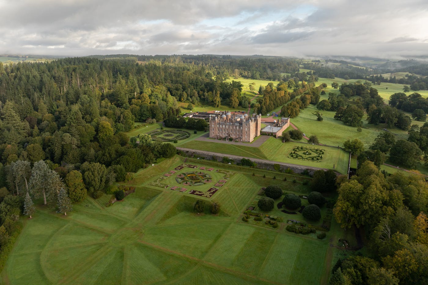 A drone view of a castle surrounded by green gardens and trees.