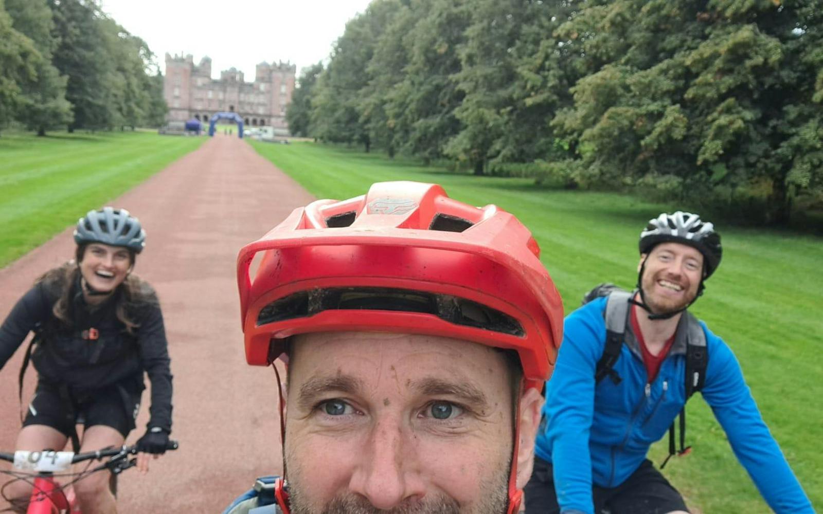 Three people taking a selfie while riding a bike in a leafy park.