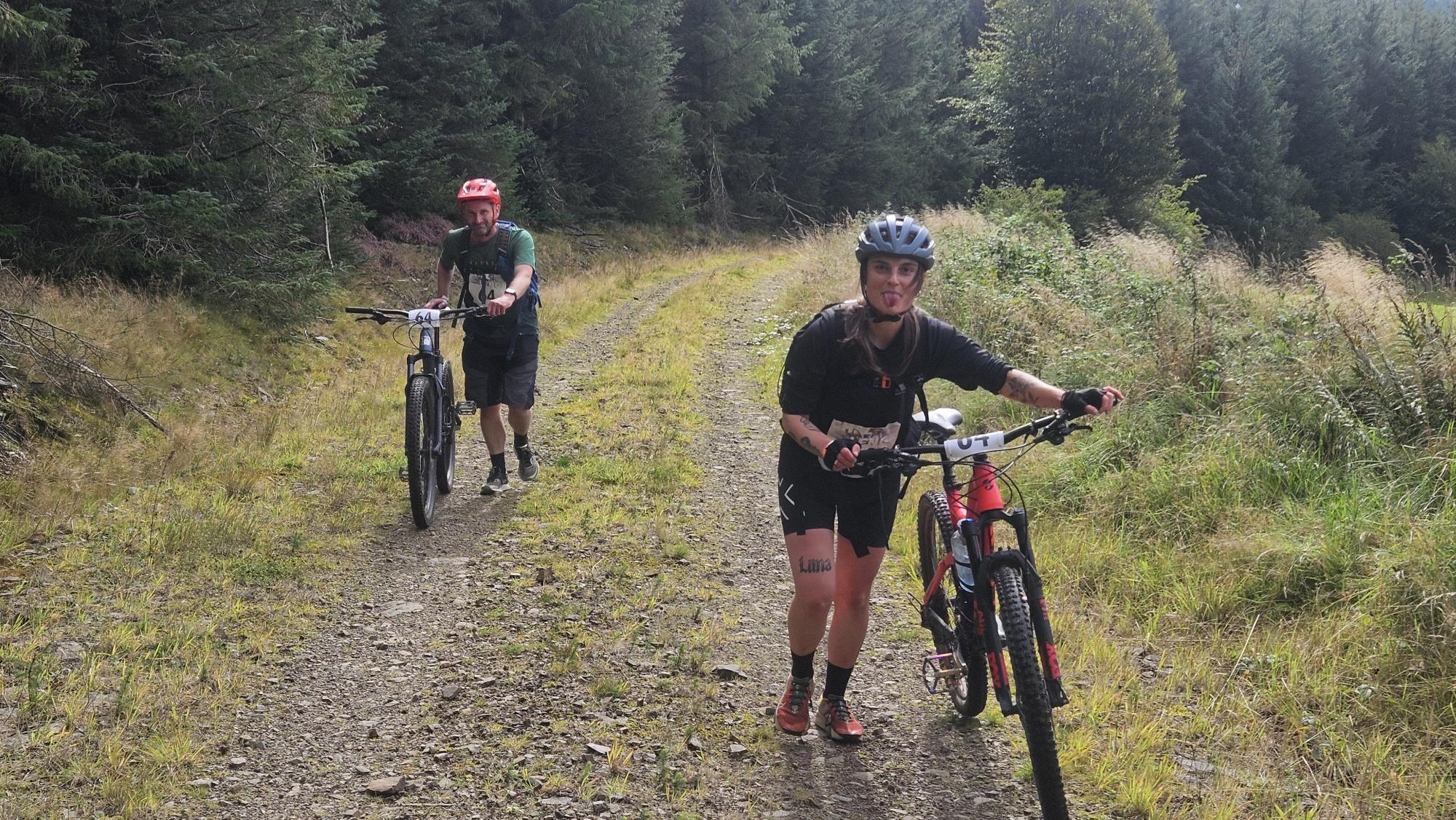 Two people in sports gear walking next to their bike in a forest.