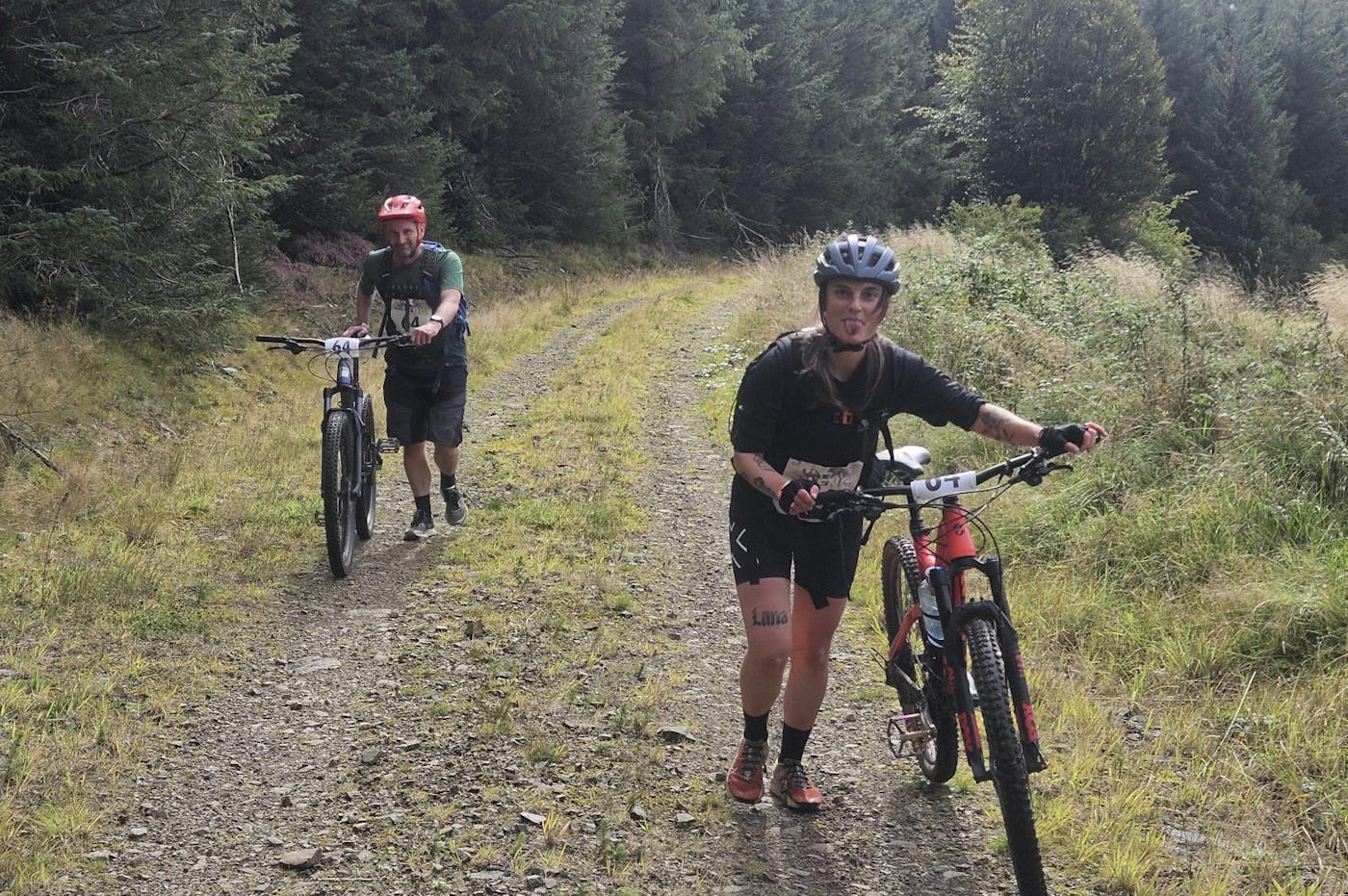 Two people in sports gear walking next to their bike in a forest.