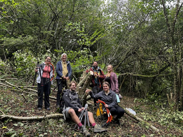 A group of six adults in a forest stand among cut branches, posing for a photo.