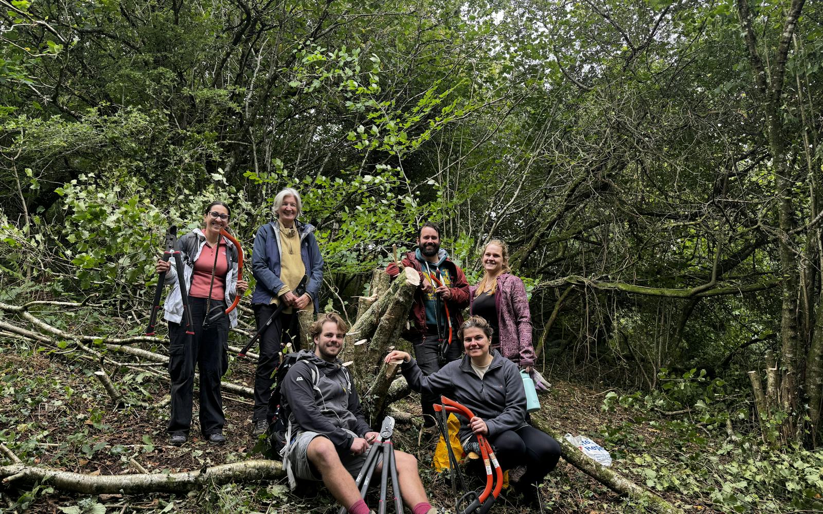 A group of six adults in a forest stand among cut branches, posing for a photo.