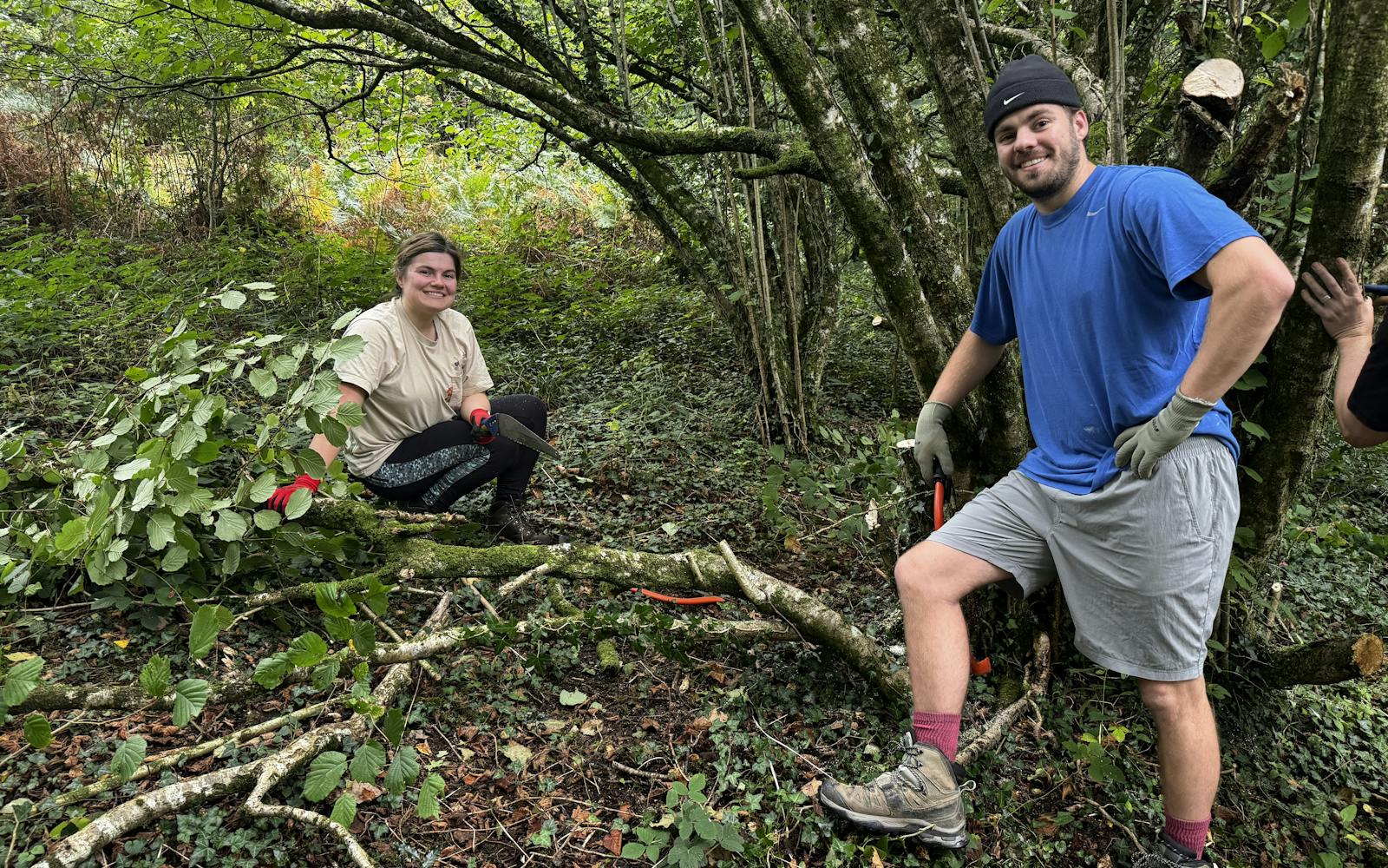 Two adults smile at the camera in a forest clearing surrounded by cut branches.