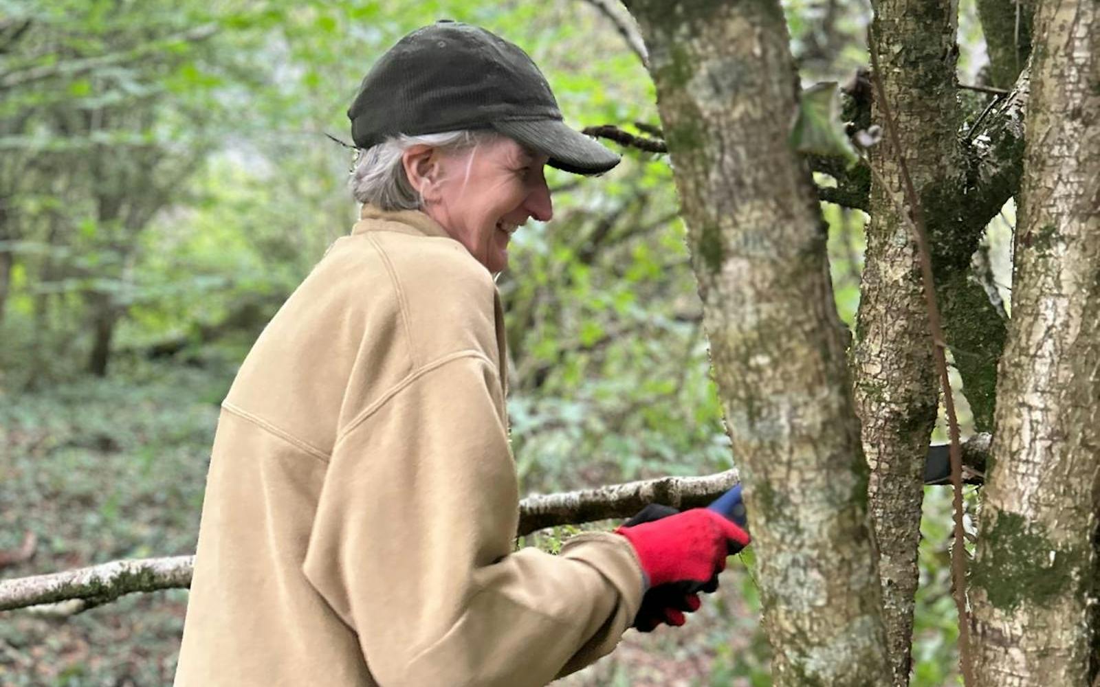 A person smiles while cutting a small tree trunk in a wooded area.