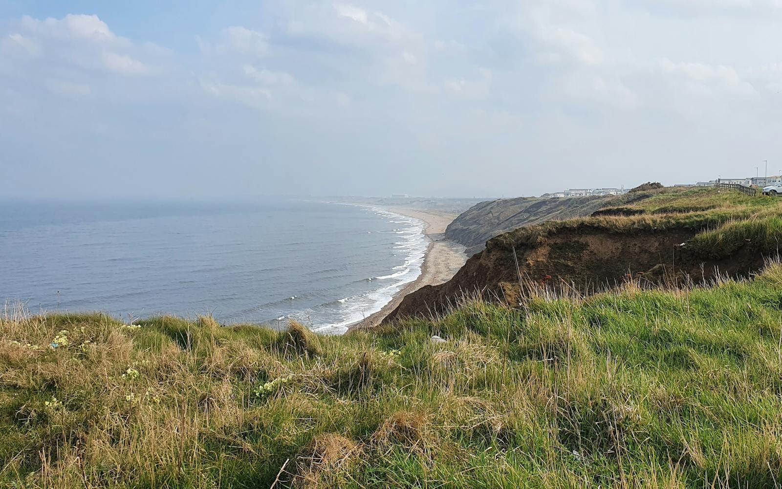 A high-angle view of a long, curving beach with crashing waves, cliffs, and a caravan park on the top right, with grassy foreground.