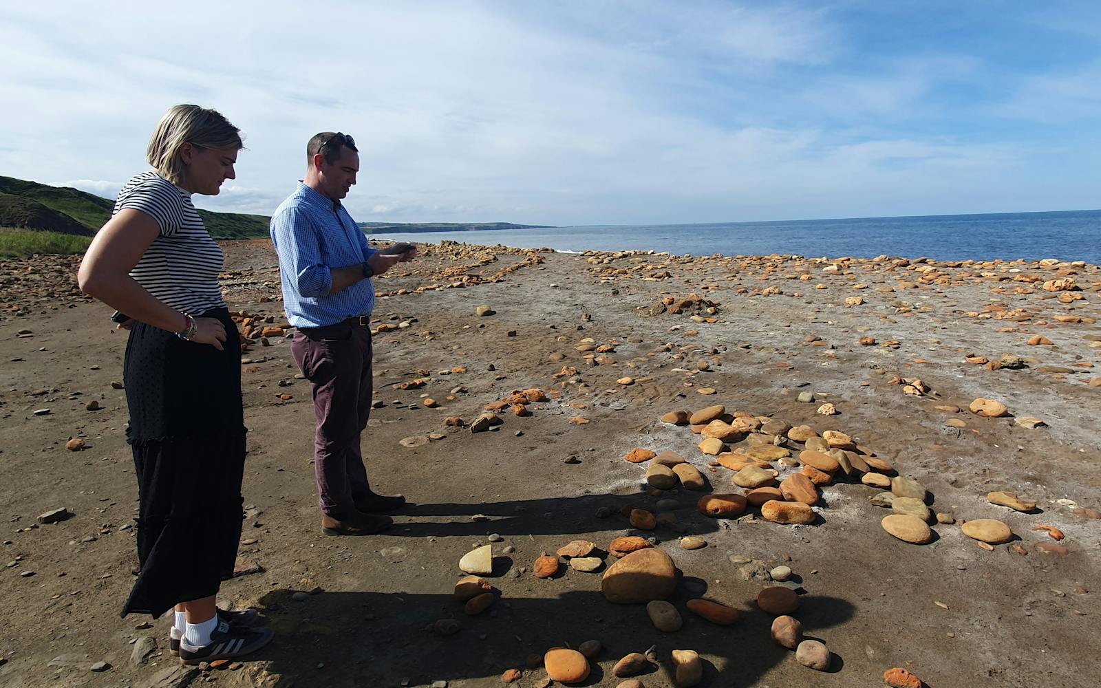 Two people, a woman and a man, stand on a dirt and stone beach, looking down at round, reddish-orange stones arranged in patterns on the ground, with the ocean and hills in the background under a blue sky.