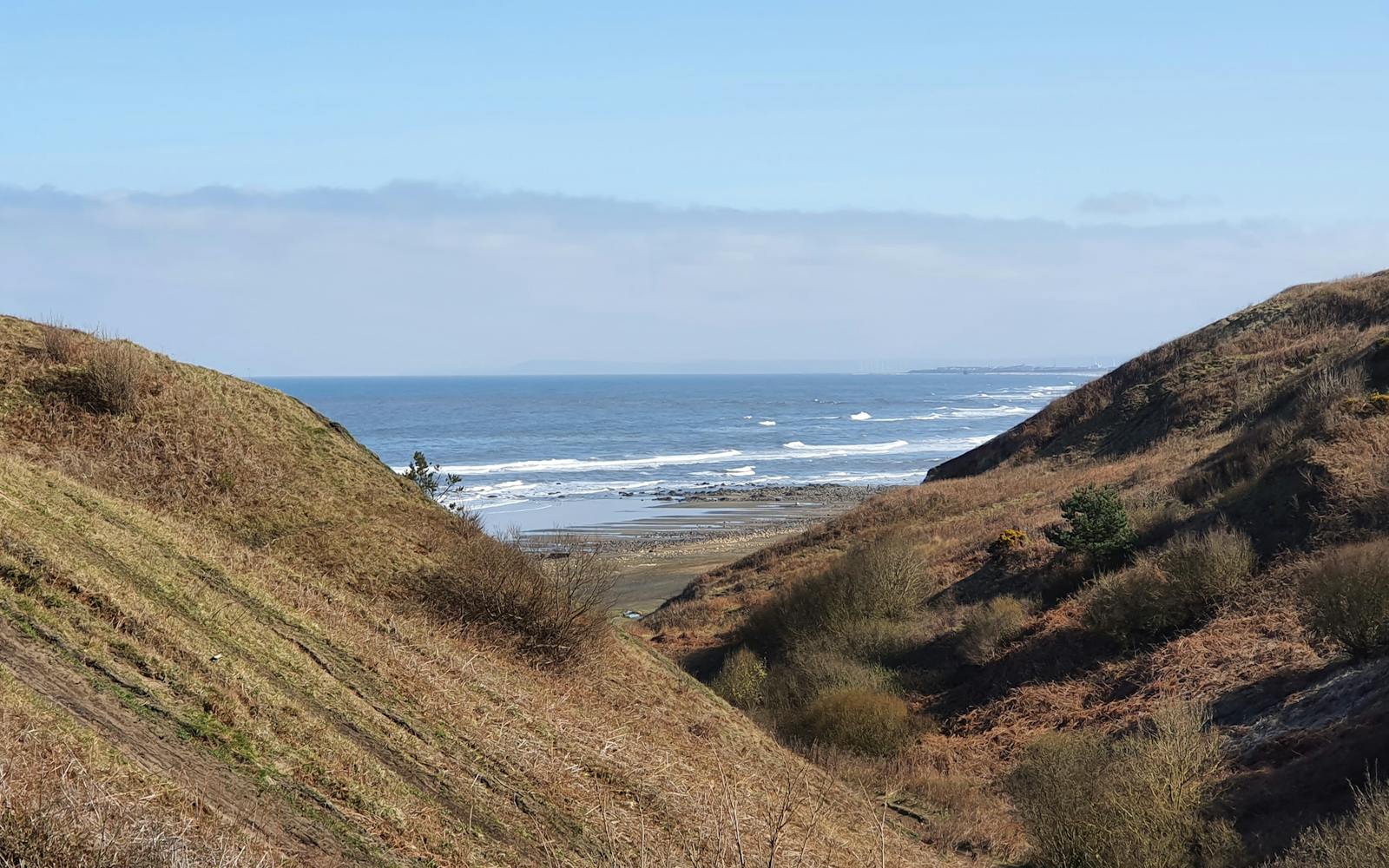 A view down a steep, grassy valley or gully toward a beach and the ocean, with waves breaking on the shore under a bright blue sky.