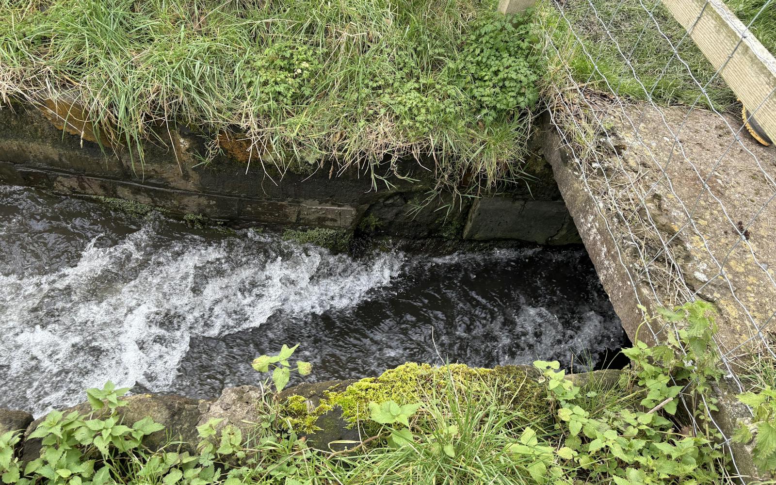 A high-angle close-up of fast-flowing water in a narrow, man-made channel or drainage ditch, bordered by concrete, grassy banks, and a wire fence and wooden post.