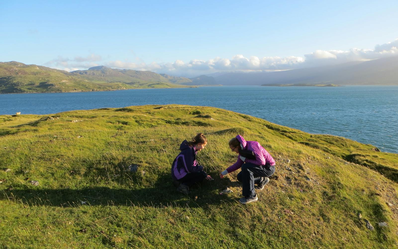 Two women seated on a green hill surrounded by a blue lake under a sunny sky.