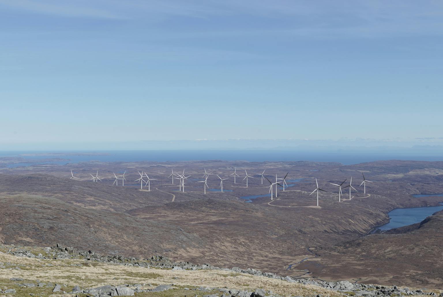 Panoramic view of a rugged coastal landscape with hills and lochs, featuring a large wind farm with numerous turbines under a clear blue sky.